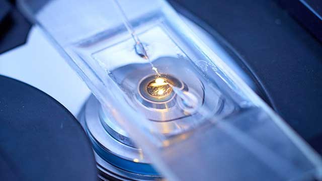 Eine Wissenschaftlerin und ein Wissenschaftler arbeiten hinter einer Glasfassade und mischen Chemikalien mit Großgeräten.